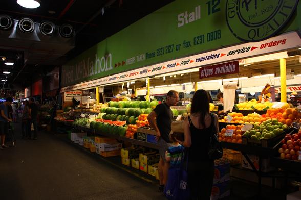 A produce display is illuminated by light in a dark store, making the fruits look colorful and inviting. The food is stacked with repeating rows. A man and woman are standing at the display having a conversation.