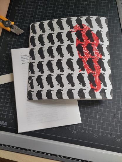 Cutting mat with on top a text block for a pamphlet with on top the paper for the cover. The cover has a block print of a red raven with a pattern of small black ravens. The fold in the cover is on the right.