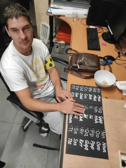 Eine Person hat beide Hände auf schwarzen Tafeln. Auf den Tafeln steht das Alphabet in Braille, Großbuchstaben, Kleinbuchstaben in Druckbuchstaben und in Schreibschrift. Die Tafeln liegen auf einem Tisch gemeinsam mit einer Tasche, einer Schüssel mit Nüssen und Computerausstattung. Die Person trägt eine gelbe Armschleife mit drei schwarzen Punkten.