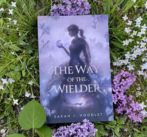 A copy of The Way of the Wielder, by Sarah J. Hoodlet, against a backdrop of green grass and spring flowers.
The cover features a young woman, standing profile, staring at a glowing orb in her hand. A misty cityscape is behind her, and the vignette (between the title and the author's name) features a fox running through a mountainous landscape.
The flowers are a combination of phlox and lilac, in shades of purple and creamy white. The grass and leaves are vibrantly green.