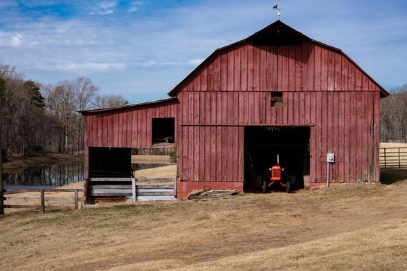 A color photo of a large red barn sitting in a field next to a pond with a forest on to opposite bank.