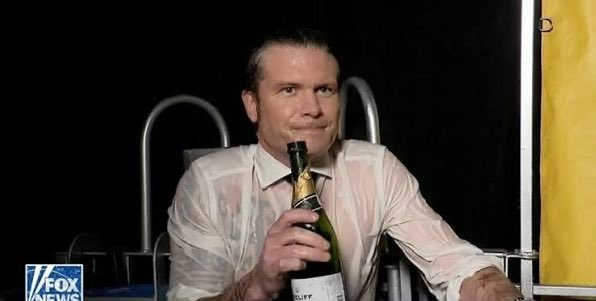 Night-time photo of Pete Hegseth in a clingy, transparent because wet white shirt & tie clutching an open Champaign bottle with what looks the the rails of a pool ladder behind him and a Fox News logo on the bottom left hand corner. The tight-lipped expression on his face suggests he's just taken a swig of fizzy champagne.