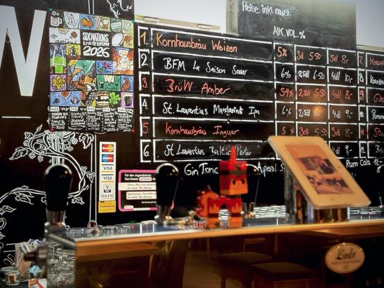 Eine Barszene mit einem Tafelmenü, das verschiedene Biere mit ihren Namen und Preisen auflistet, zusammen mit einer farbenfrohen Kalenderanzeige für eine Veranstaltung mit dem Titel "Skinations Live Event 2026". Im Vordergrund sind eine kleine dekorative Figur und ein Tablet-Ständer zu sehen,