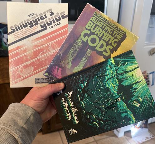 A hand holds three tabletop role-playing game books: "The Smuggler's Guide to 1365," "Awaiting the Burning Gods," and a book called “Down Among the Dead” featuring a green and black cover with a skeleton design.