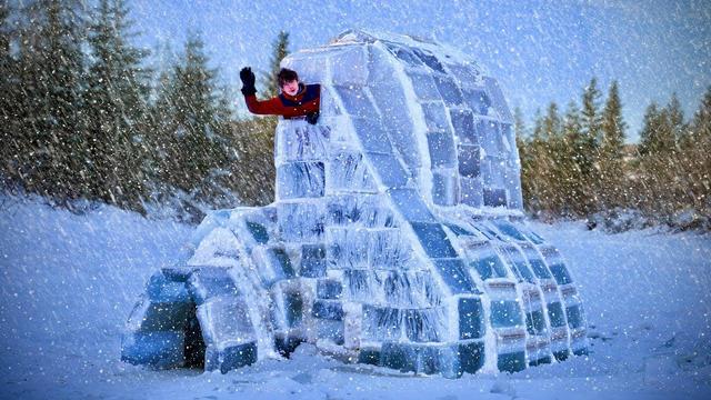 Yogoman in AK waves from the top of a two-story ice block structure built from frozen lake water in a snowy Alaskan landscape