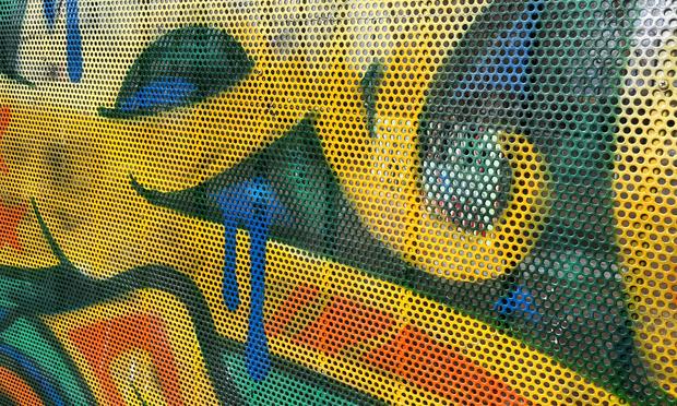 A perforated metal fence at a local skatepark spray painted with colorful, abstract swirls of yellow, orange, green, and a few drips and splatters of blue.