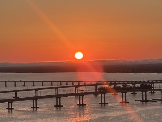 Wider River shot with island: Long low calm river with a dark strip of land in the horizon, a long unbroken strip of clouds, late afternoon. White hot sun with yellow edge and a red orange sunlight arrow. And a round lens flare. Pic by me.  Sky deeper orange red.
