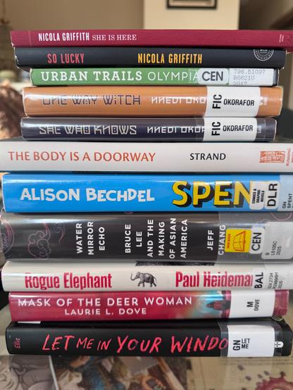A stack of books, mostly from a library, on a glass topped coffee table. Titles from bottom to top: Let Me In Your Window by Adam Ellis, Mask of the Deer Woman by Laurie L. Dove, Rogue Elephant by Paul Heideman, Water Mirror Echo by Jeff Chang, Spent by Alison Bechdel, The Body is a Doorway by Sophie Strand, She Who Knows and One Way Witch by Nnedi Okorafor, Urban Trails Olympia by Craig Romano, and So Lucky and She is Here by Nicola Griffith.
