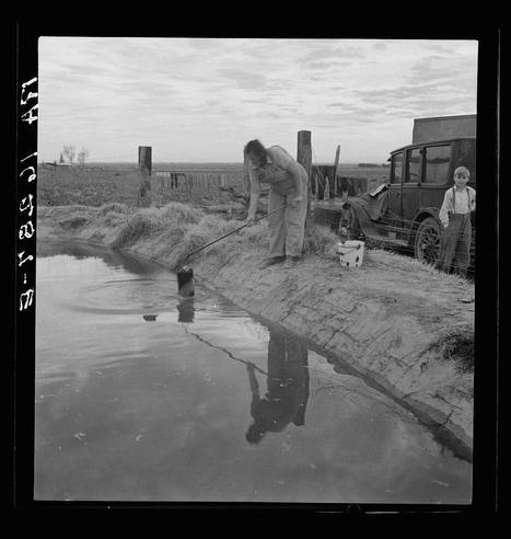 The image is a black and white photograph depicting an outdoor scene with two individuals near what appears to be an irrigation ditch. On the left side, there's an adult male leaning over the edge of the water-filled ditch, holding onto its rim as he looks into it or possibly reaching towards something within it using his right hand that holds a thin object like a stick or rod. His reflection is visible on the surface of the water.

To the far right stands a young boy, wearing trousers and shoes with laces tied at the ankles, looking toward the adult male's direction while standing close to what appears to be an old-fashioned cart or wagon attached by ropes to a wooden structure in the background. The ground around them is barren with sparse grass, indicating arid conditions.

Behind this main scene stands another older vehicle and various farm implements such as wire baskets that suggest agricultural activity nearby. In the far distance, beyond the immediate vicinity of these individuals, there's an open field or rural landscape under a partly cloudy sky. A wooden fence runs along one side of the ditch, adding to the rustic setting.

The image has several identifying marks on its edge, including what seems like handwritten text and symbols that could be part of a photographic collection cataloging system but are not fully legible due to perspective. The overall ambiance  [...]