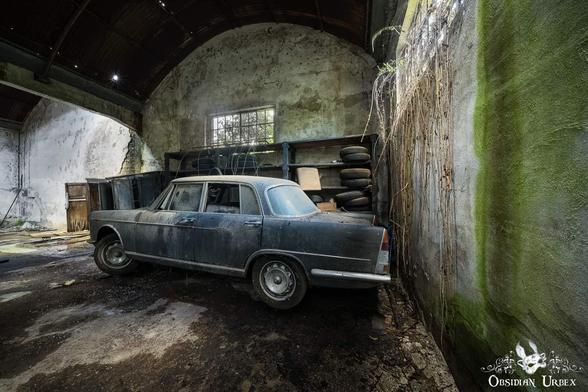 An old, dusty dark blue car is parked inside a decaying building with mossy walls and an arched ceiling. Shelves with tires are visible to the right.