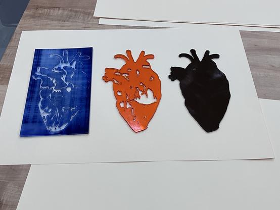 Photo of the three acrylic plates used to print the multilayer anatomical hearts in the previous photo. A rectangular acrylic sheet with lightly heart-shaped details is stained blue from ink; two heart-shaped acrylic sheets in orange and dark red acrylic are shaped to provide background shape and detail for the heart print.