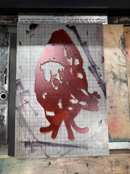 Photo of an acrylic plate used to print the multilayer anatomical hearts in the previous photos. It's a heart-shaped acrylic sheet in orange that's been inked in red,  shaped to provide midground shape and detail for the heart print. It's sitting on a silver gridded boxcar base to help bring it to type height on a press bed so it's high enough to be printed.