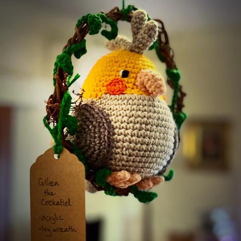 A crocheted yellow and grey cockatiel named Colleen hangs inside a small ivy-wrapped wreath. A brown tag describes the materials used: acrylic yarn and ivy