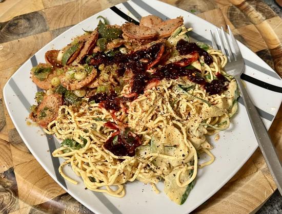 a square plate on a wooden cutting board. to the right is a fork. 
the plate is loaded with two main components. on the bottom side is thin noodles and strips of cucumber dressed in a creamy pepper yoghurt and tahini sauce. the top side of the plate has thinly sliced sausage and spring onions, with a topping of dark red beetroot relish