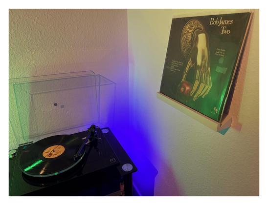 Auf einem schwarzen Plattenspieler läuft eine schwarze Schallplatte. Rechts an der Wand ist ein kleines weißes Regalbrett, darauf steht das Cover. Schwarz, darauf ist eine golden Hand, die nach unten hängt und einen roten Apfel hält.