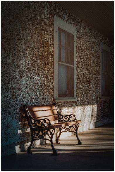 A wooden bench with ornate black metal legs stands against a textured, weathered wall with two tall windows. Warm late-afternoon sunlight falls diagonally across the bench and pavement, casting long shadows. The scene feels quiet and empty, with soft grain typical of film photography.