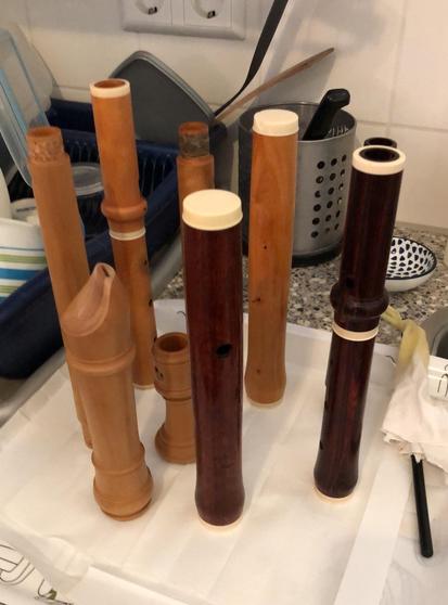 Parts of wooden flutes, in a yellowish and a darker brown wood, standing upright on a tray covered with paper towels. An oily rag attached to a stick. Some dishes on a drying rack in the background.