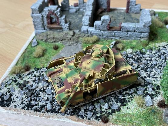 A photo of a plastic scale model of a german WW2 tank (close up on the tank). It is colored with camo marking. It is a part of a diorama from the period, comprising a ruined stone house with no roof, some low grass around it, a tree and a gravel road. The tank is in the middle of the the road. Its cannon is pointed away from the house, toward the camera.