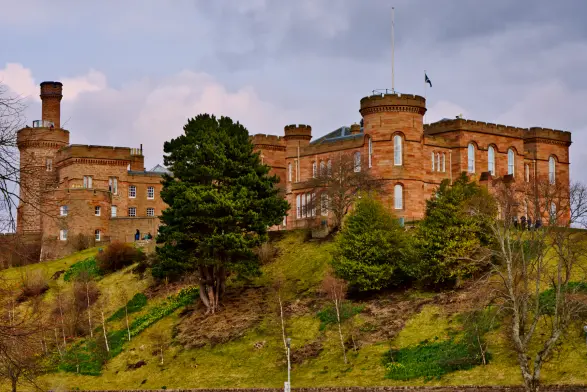 Inverness Castle mit seinem typischen roten Aussehen thront auf einem grünen Hügel. Ein großer Baum in der Bildmitte verdeckt einen kleinen Teil des Bauwerkes. Die ersten Blumen recken ihre Blüten dem Himmel entgegen. 
. 
Inverness Castle, with its characteristic red appearance, sits proudly atop a green hill. A large tree in the center of the picture partly obscures the building. The first flowers are stretching their blossoms toward the sky. 
.
