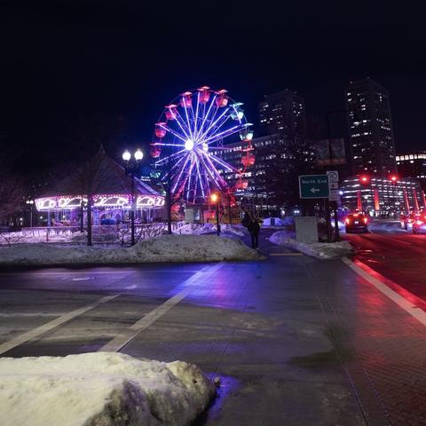 A nighttime scene featuring a brightly lit ferris wheel and carousel. Snow covers the ground, and city buildings are in the background. Streetlights and traffic signals are visible, adding to the urban atmosphere.