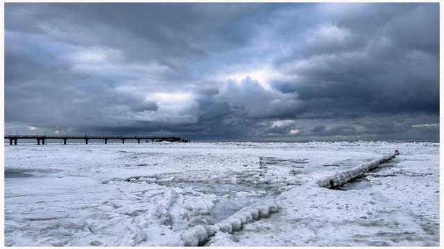 A winter landscape featuring a frozen body of water with patches of ice and snow. A wooden pier extends into the icy expanse, set against a backdrop of dramatic, overcast skies filled with dark clouds.