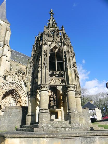 La recevresse d'Avioth, un grand monument gothique muni d'une flèche posé sur des colonnes avec au centre une statue de la Vierge à l'Enfant. A l'arrière-plan, un bout de la basilique est visible, le ciel est bleu. 
The "recevresse" of Avioth, a large Gothic monument with a spire placed on columns with a statue of the Virgin and Child in the center. In the background, part of the basilica is visible, the sky is blue.