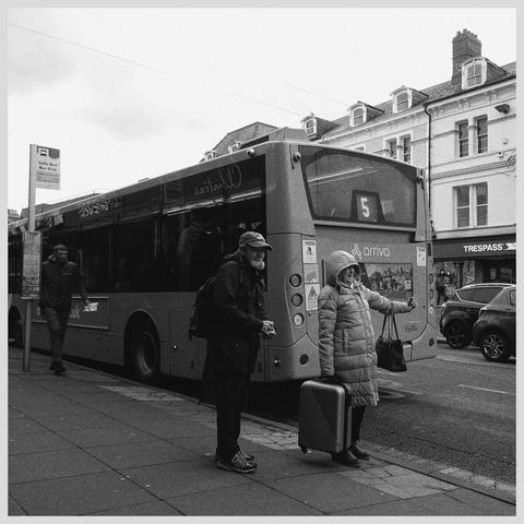 2 people waiting for the bus at the bus stop. A woman is sticking her arm out signalling for an oncoming bus to stop.