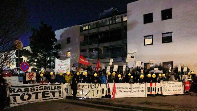 Das Bild zeigt eine abendliche Demonstration gegen eine AfD Wahlkampfveranstaltung. Eine Gruppe von „pi mal Daumen 250 Protestierenden“ (Zitat Polizei) hat sich mit Bannern und Schildern auf der Straße hinter einer Absperrung versammelt. Die Banner tragen Aufschriften wie "Rassismus tötet!", „AFD Verbot JETZT!“ sowie zwei Banner der Omas Gegen Rechts Gruppen aus Erding und Landshut mit dem Text „Null Toleranz für Intoleranz! - Demokratie hat keine Alternative!“ sowie „Gemeinsame Verantwortung für unsere Demokratie".

Die Gesichter der Menschen im Bild wurden, soweit möglicherweise erkenntbar, bearbeitet und mit einem Smiley überdeckt.