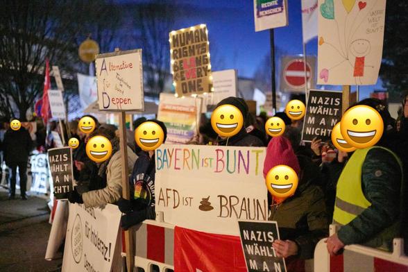 Im Vordergrund und Mittelgrund sind mehrere Protestierende zu sehen, deren Gesichter mit gelben Smiley-Emojis überdeckt sind. Sie halten verschiedene Schilder hoch, die mit handgeschriebenen Texten und Zeichnungen versehen sind.

Die Schilder tragen verschiedene Botschaften, darunter "Alternative für Deppen", "HUMAN RIGHTS BELONG EVERY ONE", "BAYERN IST BUNT", "AFD IST BRAUN" und "NAZIS WÄHLT MAN NICHT". Es gibt auch Schilder mit Regenbogenfarben und der Aufschrift "OMAS GEGEN RECHTS".