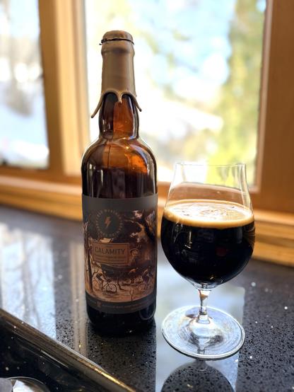 A tall brown bottle with a thick gold neck wax has a dark label showing bicycle riders pushing through a flooded field. A snifter full of the dark beer is to the right.