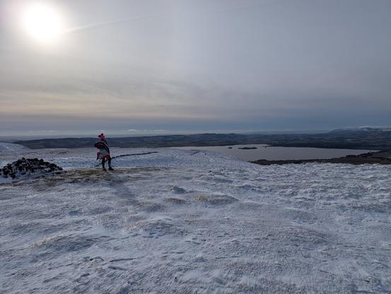 Summit of Bishops Hill a weak sun above left of a figure at the cairn. Light covering of snow and Loch Leven (Fife) behind.