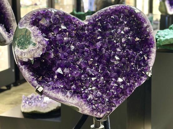 Large amethyst geode cut in half to show the cavity is filled with dark purple crystals. The outer edge of the matrix stone is gray and the left side also has a blob or patch of green malachite intruding into the heart-shaped interior. These are large crystals about 3/4” to 1” wide and come to the classic points. Marcus Budil gallery.