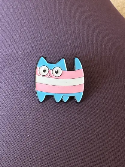 An enamel pin of a simple cartoon kitty with trans pride flag colours on its body!