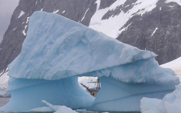 The bow of the tallship Bark Europa is viewed through a heart-shaped window in an iceberg that fills most of the image. In the background is a rocky slope on the far side of the fjord.