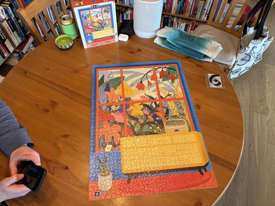 Completed jigsaw puzzle on a round wooden table. The puzzle image is an illustration of a window filled with flowers, with a radio sitting on a console table in the foreground.