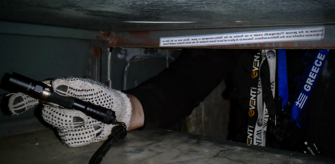 Foto im Innenraum eines alten Blechschranks. 
Aufgenommen aus dem Schrankfach heraus. 
Links im Schrankfach eine Hand mit weißem Strickhandschuh und LED-Taschenlampe. Von der Person ist nur ein Teil des Oberkörpers zu sehen vor dem Schrankfach, viele Lanyards, ua. "Greece", "Wikipedia". Ein kleines PMR-Funkgerät.
Hinter dem Türsturz des Schrankfaches klebt ein einlaminierter Zettel mit schwarzem Text auf weißem Grund. In Spiegelschrift, in Pseudo-Kyrillischer Schrift. 
"Draußen in der großen Wartungshalle scheint es ein Problem an der Elektroversorgung zu geben. Gleich hier um die Ecke im Erdgeschoss befindet sich der Elektroschaltraum. Schauen sie da gleich mal nach! Sicher können Sie das kleine Problem schnell lösen."