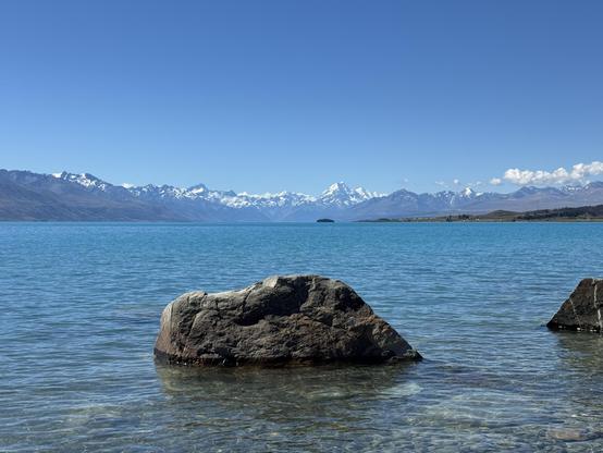 Distant view of snow topped mountains under a blue sky viewed across a long lake. A boulder emerges from the lake in the foreground