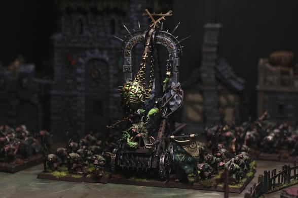 A warhammer fantasy skaven plague furnace in a dark setting, surrounded by clanrats, doomwheels, abominations and more. Its paint scheme is moody, dark with sharp highlights of metallic bronze and iron. Greys, browns and greens dominate the model.