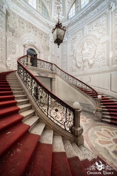 A grand, ornate staircase with a sweeping red carpet and detailed black wrought-iron railings. Intricate white wall carvings and a large chandelier adorn the elegant building.