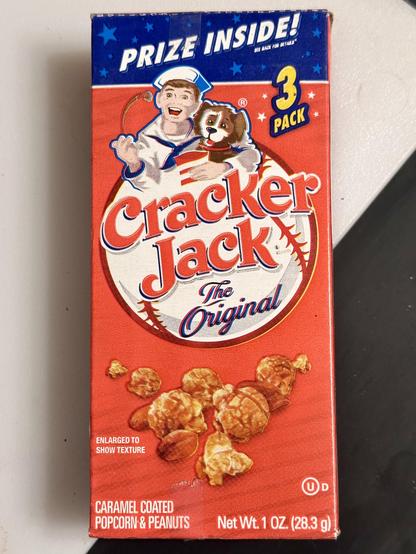 Front of familiar orange box of classic Cracker Jack, an American snack popular at baseball games back in the Day. Boy with spaniel dog cartoon are at top of baseball for orange logo with name and “The Original” below. Near bottom is a photo of caramel corn and peanuts. Fine pint says “enlarged to show texture” but the product inside is twice this size.