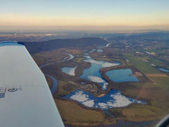 Look out of a light aircraft with parts of the wing in view. A group of small turquoise/blue lakes, partly covered with floating ice. A small range of hills in the background under a blue sky with light clouds near the horizon, lit orange from a warm evening sun. The airfield is visible at the right edge of the frame.