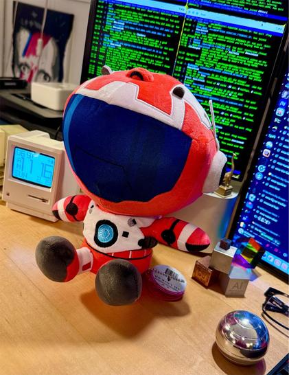 No Man's Sky Traveller Plush from YouTooz sitting on a desk in front of a computer setup displaying terminal windows onscreen