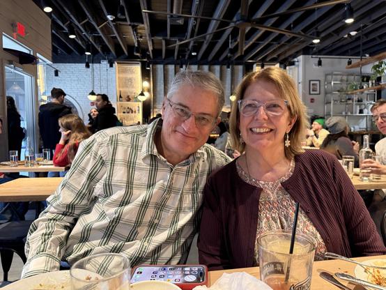 My wife and I pose for a photo at a restaurant, surrounded by other patrons. I'm wearing a plaid shirt and glasses, while Gabrielle has on a burgundy cardigan and is holding a drink. Tables are set with dishes and drinks.