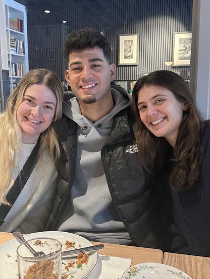 A group of three young adults is smiling together at a dining table. Two women are on the left and right, while a man sits in the center. Plates with leftover food and drinks are on the table. The background features bookshelves and framed artwork. My youngest daughter Adrienne is on the left, my oldest daughter Rhiannon is on the right, and her fiancee Chris is in the middle.