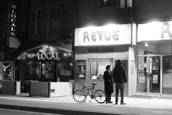 Night outside the Revue Cinema and The Local pub. Their lights illuminate a couple standing on the street beside a bike chained to a bike stand.