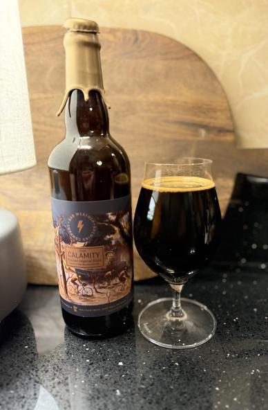 A tall brown bottle with a golden wax neck ring has a label in shades of brown, bicyclists mired in a swamp try to traverse the mess. A snifter glass full of beer is to the right.