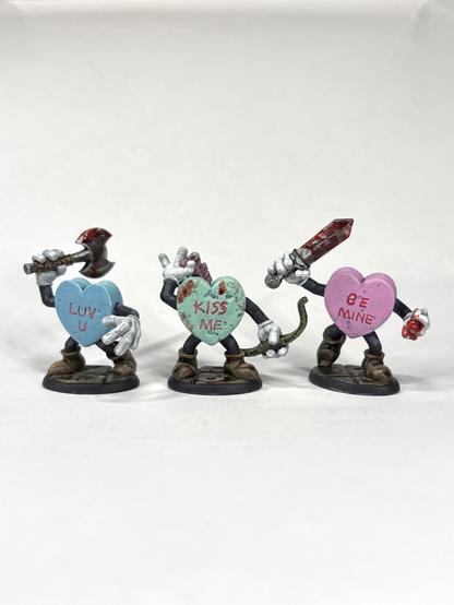 Three candy hearts with arms, legs, and weapons. They are moving aggressively forward. “LUV U”, “KISS ME”, and “BE MINE” are written in them. They are light blue, sea green, and pink and have blood splattered on them.