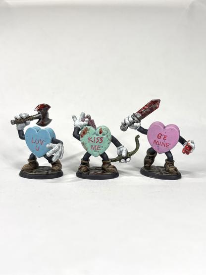 Three candy hearts with arms, legs, and weapons. They are moving aggressively forward. “LUV U”, “KISS ME”, and “BE MINE” are written in them. They are light blue, sea green, and pink and have blood splattered on them.