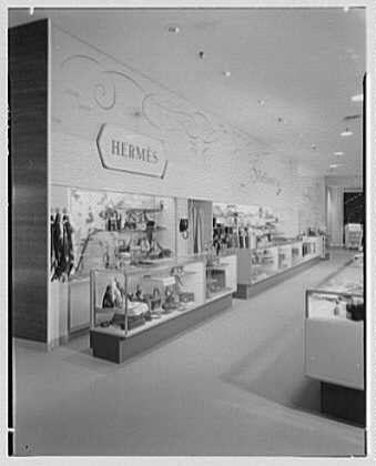 The image depicts an old department store, specifically the interior of a luxury goods section for the brand Hermes. The photograph is in black and white, giving it a vintage feel consistent with its March 6th, 1956 date mentioned on the negative caption.
Several display cases line one side of the aisle showcasing various items such as watches, sunglasses, handbags, leather goods, and possibly jewelry pieces. The merchandise displays are neatly arranged in glass cabinets that allow customers to view the products from multiple angles without obstruction.
Prominently displayed above these showcases is a large sign with "Hermes" written on it, indicating the brand being featured at this particular section of the store. To the left side of the aisle, additional clothing items hang near the entrance area, suggesting that there might be more merchandise just outside of what's visible in this photograph.
The ceiling features decorative light fixtures and a network of curved lines or perhaps wiring, adding to the overall elegance of the space. The flooring is not clearly discernible but appears to have a clean, polished look appropriate for a high-end retail environment. Overall, the store exudes an air of sophistication and exclusivity typical of luxury department stores during that era.
This image provides insight into how brand-specific areas were presented in mid-20th-century [...]