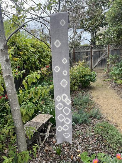 A purpleish-gray scarf with white irregular rings hanging from the branch of a tree in a garden.